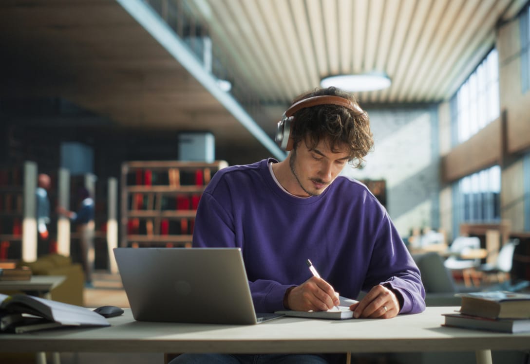 joven con cascos estudia en la biblioteca con apuntes y ordenador.
