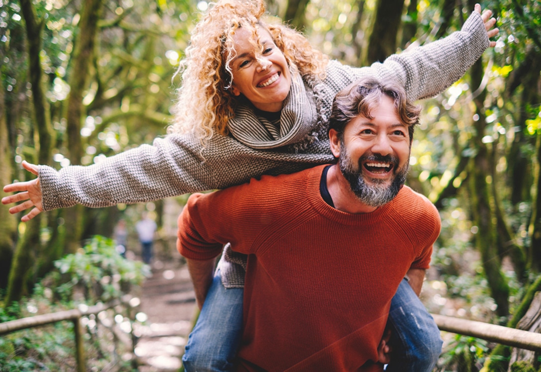 pareja-hormona-felicidad hombre carga sobre la espalda a mujer en entorno natural, ambos sonries y ella estira los brazos