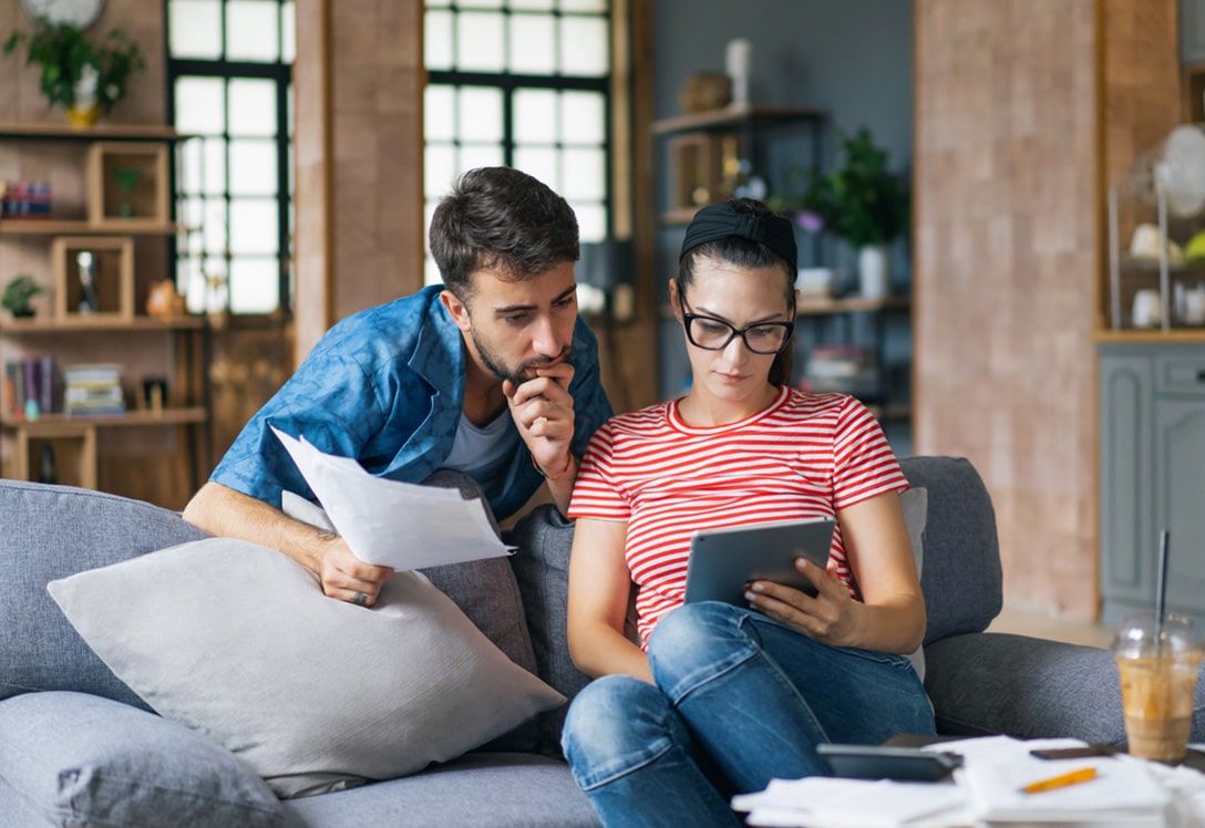 pareja-joven-ayudas-2026 pareja haciendo cuentas y buscando ayudas para jóvenes en el salón de casa.